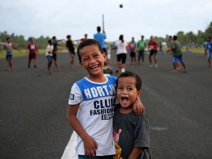 Two kids pose for the photo while a volleyball game is played on the Funafuti airstrip in the background