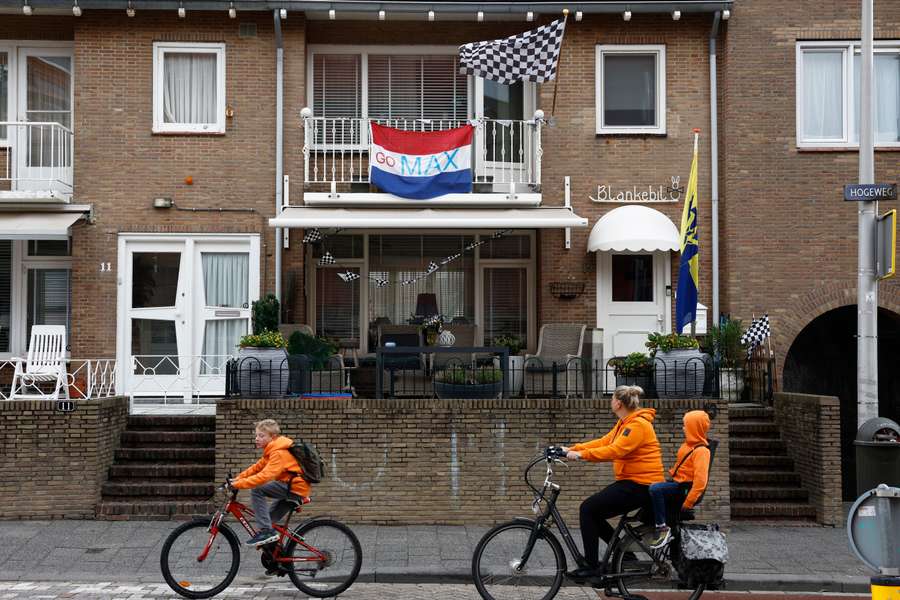 Ciclistas vestidos de laranja passam por uma casa onde se lê uma bandeira holandesa 
