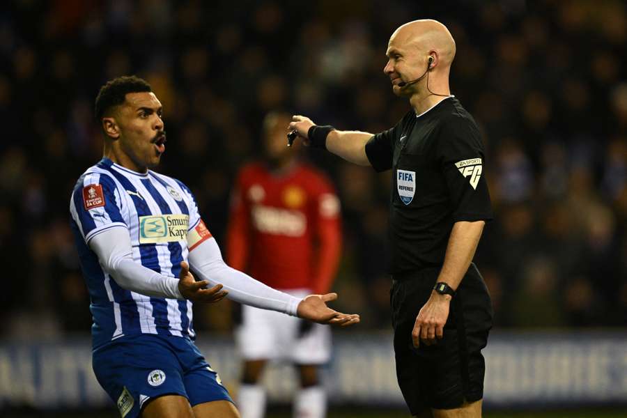 L'attaccante nordirlandese del Wigan n. 28 Josh Magennis (L) reagisce mentre l'arbitro inglese Anthony Taylor (R) assegna un rigore allo United