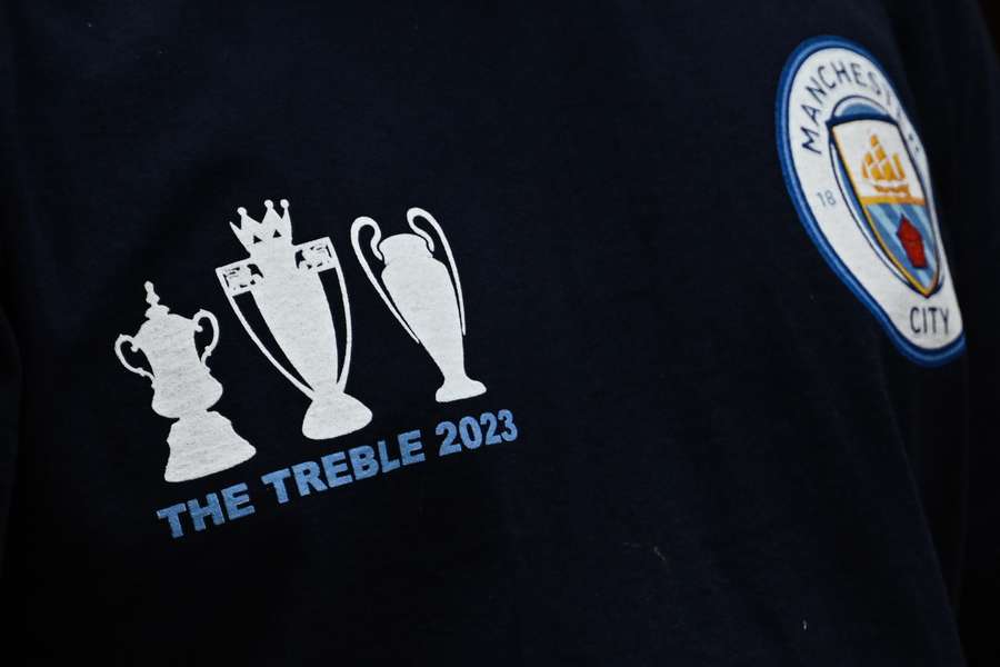 A Man City fan wears a t-shirt featuring all three trophies